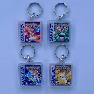 May include: Four clear acrylic keyrings featuring Pokémon Game Boy game covers. Each keyring has a silver ring and displays a different game version: Red, Green, Blue, and Yellow. The text "GAME BOY" and "Pokémon" are visible on each keyring.