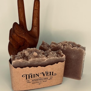 Op de afbeelding: Twee stukken botanische zeep met een textuur, naast een houten handsculptuur in een vredesteken. De zeep zit in een bruine papieren hoes met de tekst "Thin Veil Apothecary Botanical Soap". De zeep is lichtbruin.