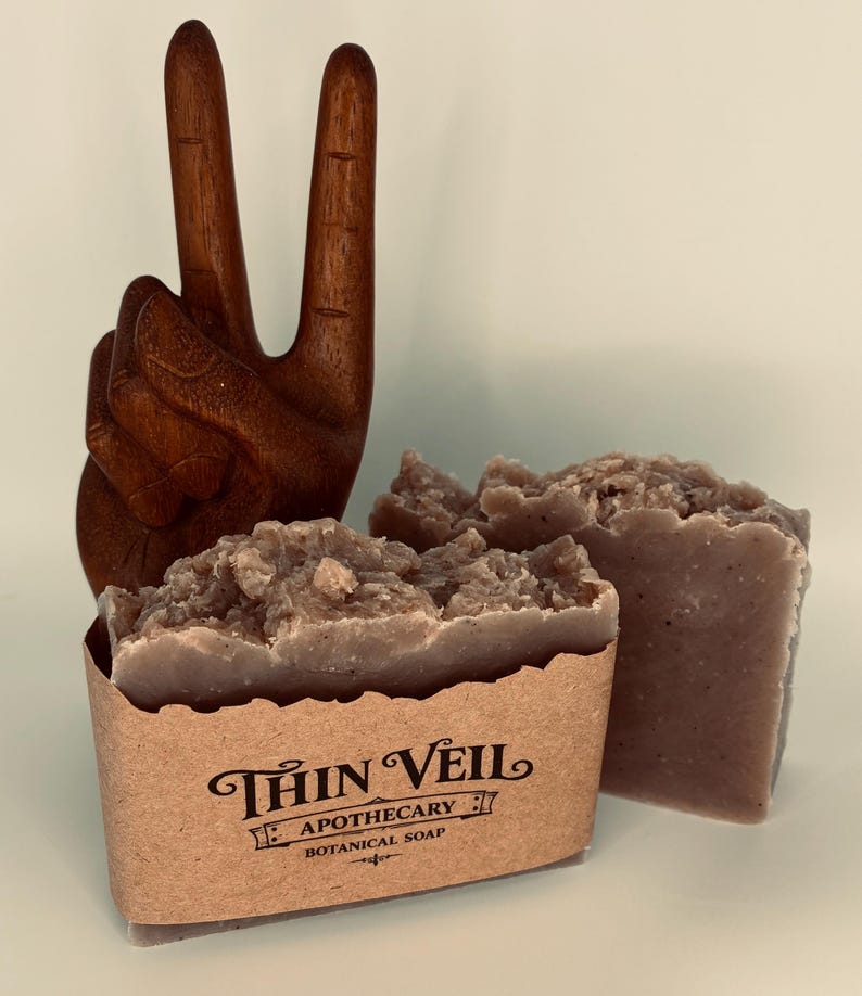May include: Two bars of botanical soap with a textured surface, next to a wooden hand sculpture in a peace sign. The soap is in a brown paper sleeve with the text "Thin Veil Apothecary Botanical Soap". The soap is a light brown colour.