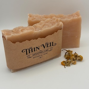 Puede incluir: Dos barras de jabón botánico de color melocotón claro. Una barra está parcialmente envuelta en una etiqueta de papel marrón con el texto "Thin Veil Apothecary Botanical Soap". Flores de manzanilla secas están dispersas cerca. El jabón tiene una superficie texturizada.