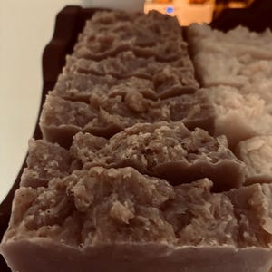 May include: Close-up of several bars of handmade soap. The soap has a textured, rustic appearance with a light brown colour. The bars are rectangular and appear to be freshly cut, with a slightly uneven surface. The soap is arranged on a wooden tray.