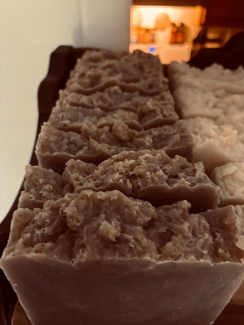 May include: Close-up of several bars of handmade soap. The soap has a textured, rustic appearance with a light brown colour. The bars are rectangular and appear to be freshly cut, with a slightly uneven surface. The soap is arranged on a wooden tray.