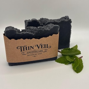 Op de afbeelding: Twee donkergrijze botanische zeepblokken, waarvan één verpakt in een bruin papieren etiket met de tekst "Thin Veil Apothecary Botanical Soap". Naast de zeep liggen verse groene muntbladeren. De zeep heeft een textuur, ongelijkmatig oppervlak.
