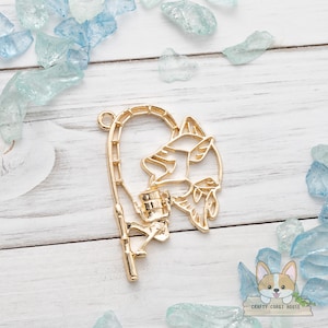 Può includere: Un ciondolo in metallo color oro raffigurante un cane corgi che pesca. Il ciondolo ha la forma di una canna da pesca con la sagoma di un cane corgi e un pesce. Il ciondolo è circondato da pezzi di vetro decorativi blu e trasparenti.