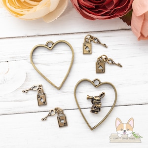 Puede incluir: Cuatro encantos de metal dorado con un marco en forma de corazón, una llave y un candado con un as de picas. Un encanto presenta un marco en forma de corazón con una figura en miniatura del Conejo Blanco de Alicia en el País de las Maravillas.