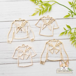 Puede incluir: Cuatro dijes de metal dorado en forma de uniformes de marinero con corbata de lazo. Los dijes están sobre un fondo de madera blanca.