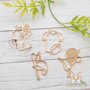 Puede incluir: Cinco dijes de metal dorado con diseños de temática deportiva. Los dijes incluyen un bate de béisbol y una pelota, una raqueta de tenis y una pelota, un balón de fútbol americano y una camiseta, y dos camisetas. Los dijes están sobre un fondo de madera blanco.