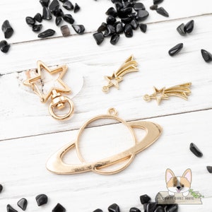 Puede incluir: Encantos de metal dorado en forma de estrella, estrella fugaz y planeta con anillos. Los encantos están sobre una superficie de madera blanca con piedras preciosas negras esparcidas alrededor.