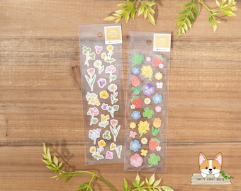 May include: Two sheets of clear sticker paper with floral and clover designs. The stickers are on a wooden background. The text on the sticker sheets reads "NB Golden" and "NB Clover".