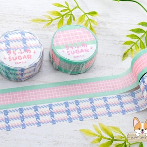 Pode incluir: Dois rolos de fita washi com um padr&atilde;o xadrez rosa e verde. Um rolo tem o texto "Sugar" em letras rosa sobre um fundo verde. O outro rolo tem o texto "Sugar" em letras brancas sobre um fundo azul. A fita est&aacute; sobre uma superf&iacute;cie branca com folhas verdes.