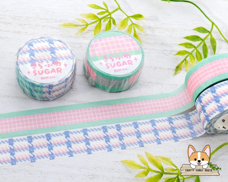 Pode incluir: Dois rolos de fita washi com um padr&atilde;o xadrez rosa e verde. Um rolo tem o texto "Sugar" em letras rosa sobre um fundo verde. O outro rolo tem o texto "Sugar" em letras brancas sobre um fundo azul. A fita est&aacute; sobre uma superf&iacute;cie branca com folhas verdes.