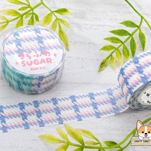 Pode incluir: Um rolo de fita washi com padr&atilde;o azul e rosa com o texto "Sugar" e "BGM Inc." impresso no rolo. A fita est&aacute; sobre uma superf&iacute;cie branca com folhas verdes.