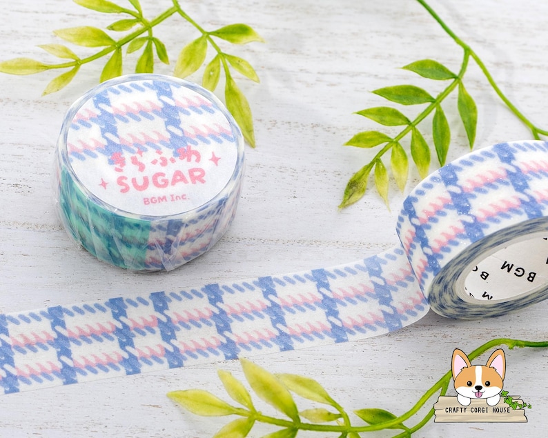 Pode incluir: Um rolo de fita washi com padr&atilde;o azul e rosa com o texto "Sugar" e "BGM Inc." impresso no rolo. A fita est&aacute; sobre uma superf&iacute;cie branca com folhas verdes.