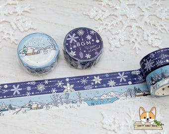 May include: Decorative washi tape rolls with winter-themed designs. One roll features a snowy landscape with houses and a snowman, while others display snowflakes. The tape is in shades of blue and white, perfect for crafts and scrapbooking.