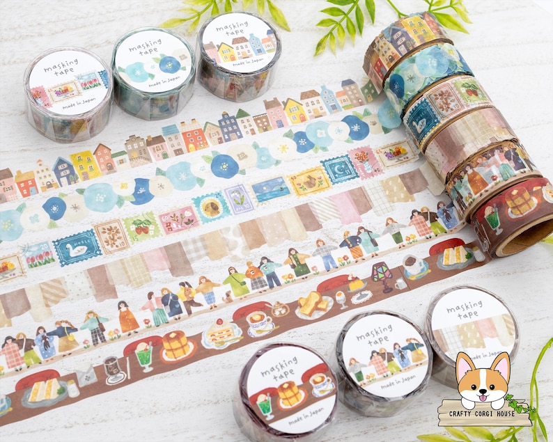 May include: Decorative masking tape rolls with various designs, including houses, flowers, and people. The tape features a variety of colors, including blue, brown, and beige. The image also shows several rolls of tape stacked together.
