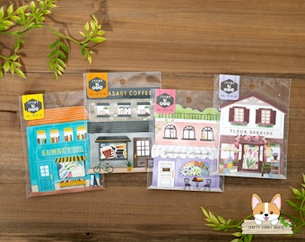 May include: Four colorful illustrated storefront stickers with Japanese text. The stickers feature a book store, a coffee shop, a bakery, and a flower shop. The stickers are packaged in clear plastic sleeves.