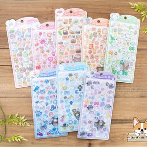 Puede incluir: Un conjunto de diez hojas de pegatinas de plástico transparente y de colores con varios diseños de animales y comida. Las pegatinas están dispuestas en dos filas de cinco hojas. Cada hoja tiene el texto "3D Pop Seal" impreso en ella.