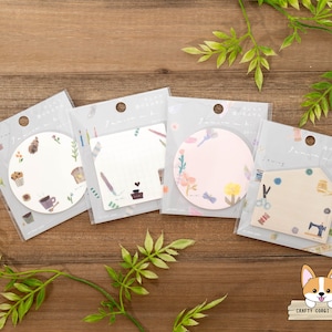 May include: Four different designs of decorative memo pads with a circle, square, and house shape. Each pad has a different design with a floral, coffee, and sewing theme. The memo pads are on a wooden surface.