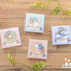 May include: Four square notepads with illustrations of storybook scenes, each in a clear plastic wrapper. The pads feature images of characters and settings from classic tales, with text in Japanese. The pads are on a wooden surface with green leaves.