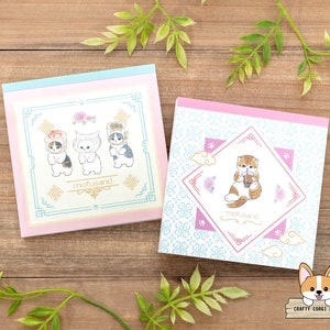 May include: Two pink and blue memo pads featuring a cute cat design. The top pad shows three cats wearing hats and the bottom pad shows a cat holding a boba tea. Both pads have the text "mofusand" printed on them.