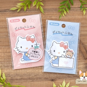 May include: Two packs of Hello Kitty themed sticky notes. The pink pack features Hello Kitty holding an apple with the text "Refrigerator Apple Pie" in Japanese. The blue pack features Hello Kitty holding a letter with a small brown bear peeking out from behind her. Both packs have the text "Die-cut Memo" in Japanese.