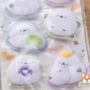 May include: A sheet of stickers featuring cartoon seals with various designs. The seals are white with gray accents and have different accessories, including a kiwi slice and a purple heart. The stickers have a light blue, yellow, and pink border.
