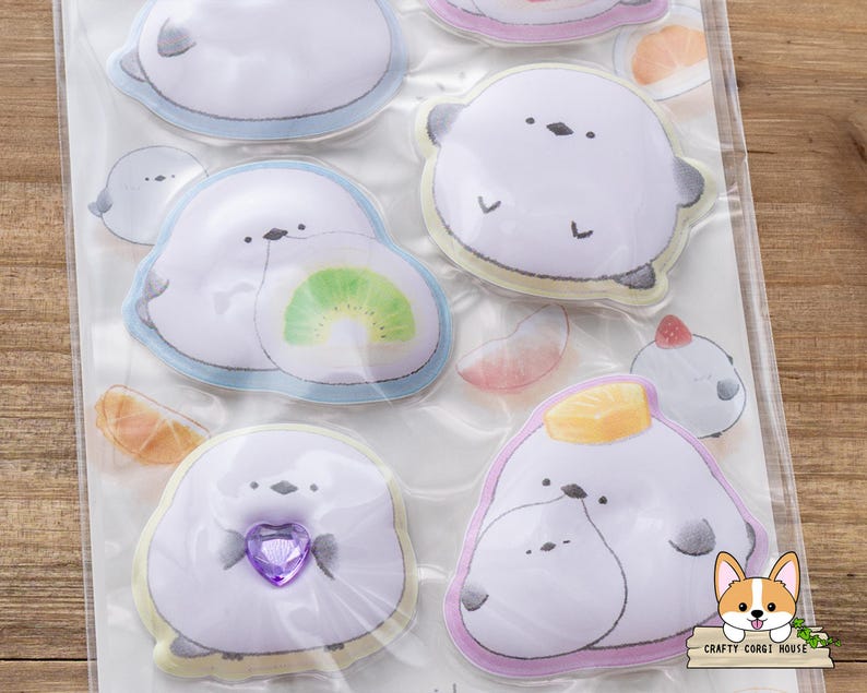 May include: A sheet of stickers featuring cartoon seals with various designs. The seals are white with gray accents and have different accessories, including a kiwi slice and a purple heart. The stickers have a light blue, yellow, and pink border.