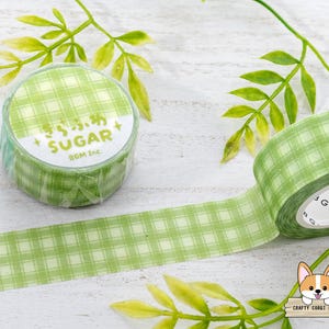 Pode incluir: Fita washi quadriculada verde e branca com o texto "Sugar" e "BGM Inc." impresso no rolo. A fita est&aacute; sobre uma superf&iacute;cie branca com folhas verdes.