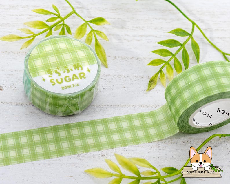 Pode incluir: Fita washi quadriculada verde e branca com o texto "Sugar" e "BGM Inc." impresso no rolo. A fita est&aacute; sobre uma superf&iacute;cie branca com folhas verdes.
