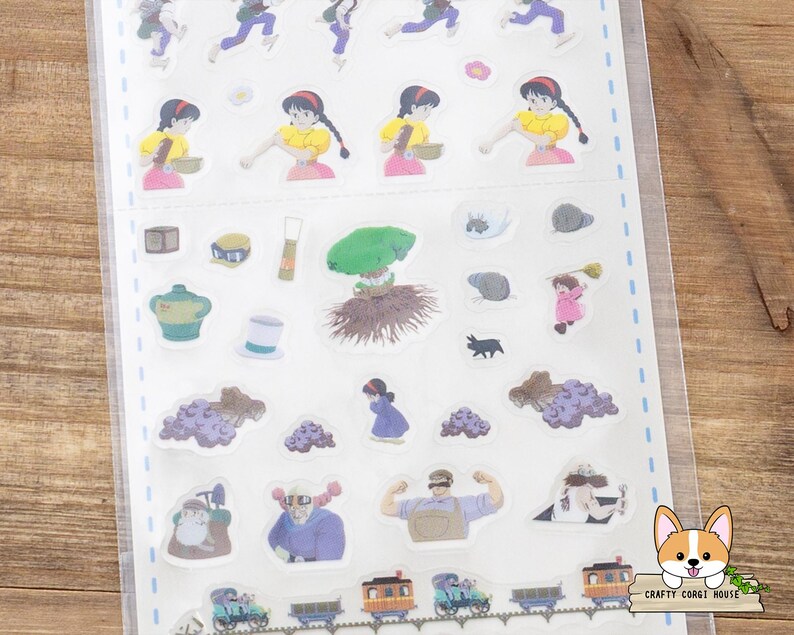 May include: A sheet of colorful stickers featuring various illustrations. The stickers include images of a girl in a yellow dress, a green tree, a black cat, and a train. The stickers are in a clear plastic sheet.