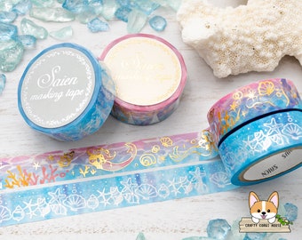 May include: Several rolls of decorative masking tape with ocean-themed designs. The tape features seashells, starfish, and coral in shades of blue, pink, and gold. The text "Saien masking tape" is visible on the tape labels. The background includes sea glass and a piece of coral.