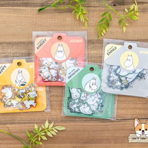 以下が含まれることがあります： 透明なプラスチックパッケージに入った4つのムーミンステッカーパック。それぞれ異なる色のラベルが付いています。ラベルには、ムーミンのキャラクターと「Happy」、「Angry」、「Thinking」、「Ennui」などの言葉が描かれています。