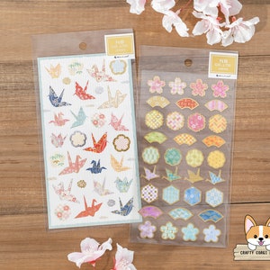 May include: Two sheets of decorative stickers. One sheet features origami cranes in various colors and patterns. The other sheet features a variety of gold, pink, and blue floral and geometric designs. The text on the sheets reads "NB CO., LTD. JAPAN".