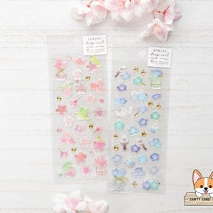 May include: Two vertical sheets of clear stickers labelled Spring drop seal. The left sheet features pink cherry blossoms and small green birds. The right sheet displays blue flowers, white birds, and gold star accents. Each sheet has a white header tag.