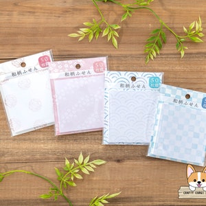 May include: Four packs of Japanese-style sticky notes with various patterns, including floral and wave designs. Each pack contains 50 sheets and is sealed in clear plastic. The background is a wooden surface with green foliage.