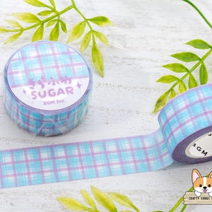 Pode incluir: Um rolo de fita washi xadrez azul e roxo com as palavras "Sugar BOM INC." impressas no rolo. A fita est&aacute; sobre uma superf&iacute;cie de madeira branca com folhas verdes.