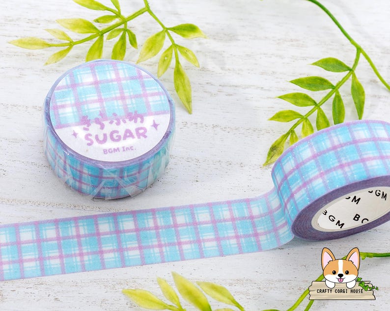 Pode incluir: Um rolo de fita washi xadrez azul e roxo com as palavras "Sugar BOM INC." impressas no rolo. A fita est&aacute; sobre uma superf&iacute;cie de madeira branca com folhas verdes.