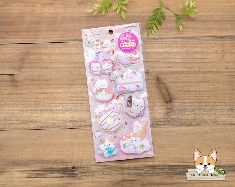 May include: A sheet of stickers featuring cartoon cats in various poses, including holding desserts and sitting on clouds. The stickers are in pastel colors with a pink background and are packaged in a clear plastic sleeve.
