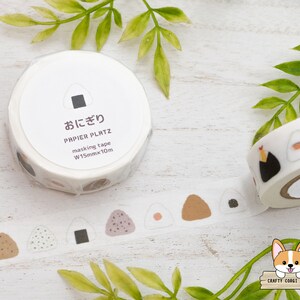 May include: A roll of white washi tape with a repeating pattern of brown, white, and black onigiri (Japanese rice balls). The tape is labeled "おにぎり" (onigiri) and "PAPIER PLATZ masking tape W15mmx10m".