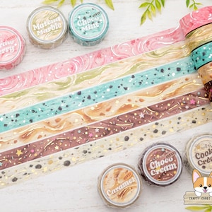 May include: A collection of decorative washi tape rolls with various designs and colors, including pink, green, brown, and gold. The tape rolls are labeled with flavors like "Strawberry", "Matcha Marble", and "Choco Mint".