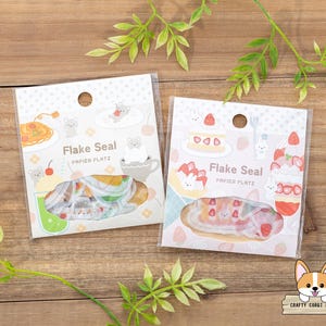 May include: Two packs of decorative paper flakes with a pastel colour scheme. The packs feature a variety of designs, including cakes, animals, and food. The text on the packs reads "Flake Seal Papier Platz".