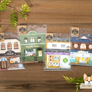 May include: Four Japanese-style paper cutouts of buildings in clear plastic packaging. The buildings include a cafe, a tailor shop, a sushi restaurant, and a traditional Japanese house. The buildings are brown, green, white, and blue.