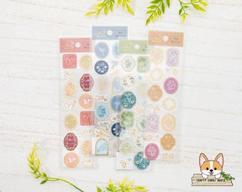 May include: Three sheets of clear sticker paper with various floral and bird designs. The stickers are in shades of blue, green, pink, and yellow. The stickers are arranged in rows and columns on the sheets. The sheets are labeled with the words "Make a Wish" and "Crafty Corgi House".