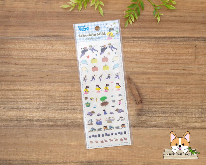May include: A clear plastic package containing a sheet of colorful stickers with the text "Schedule SEAL". The stickers feature various illustrations of people, pumpkins, and other small objects. The package is on a wooden surface.