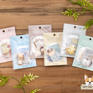 May include: Seven packs of clear flake seal stickers, each with 42 pieces, are arranged on a wooden surface. The packages are in pastel colours and branded "MOOTASTY". The stickers inside feature food-themed designs, perfect for scrapbooking or crafts.