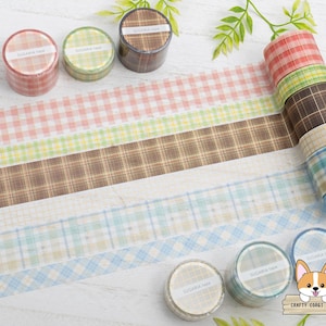 Può includere: Una collezione di rotoli di nastro washi decorativo in vari colori e motivi a quadri. I rotoli includono sfumature di rosa, verde, marrone e blu. Il nastro è srotolato per mostrare i diversi motivi. Il testo "SUGARIA tape" è visibile sui rotoli.