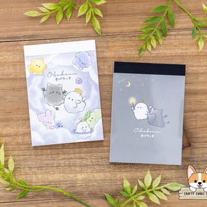 May include: Two small notepads with cartoon ghost and cat illustrations. One notepad has a white top and a light purple background with cartoon characters. The other notepad has a black top and a gray background with cartoon characters.