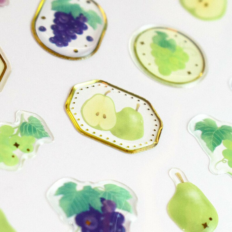 May include: A sheet of clear stickers with a gold border featuring various fruits, including grapes, pears, and apples. The stickers are perfect for adding a touch of whimsy to journals, planners, and other craft projects.