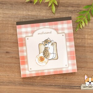 May include: A square notepad with a pink and white checked pattern. The notepad features an illustration of a cat on toast with a fried egg. The text "mofusand" is above the cat illustration.