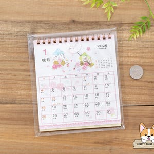 May include: A 2026 calendar with a spiral binding, encased in a clear plastic sleeve. The calendar features a pink and white design with illustrations of birds and flowers. The month of January is displayed, with Japanese characters and dates.